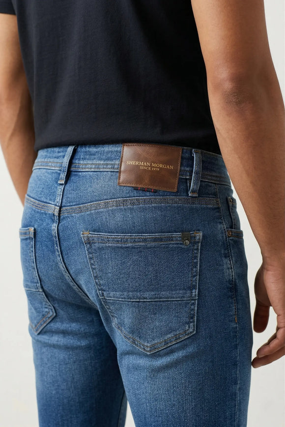 Vista posterior de jeans para hombre con etiqueta de marca en vinipiel café, placa metálica distintiva y bolsillos con diseño de franja horizontal.