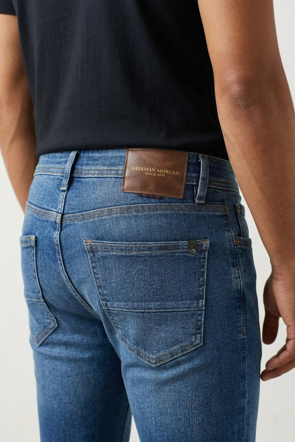 Vista posterior de jeans para hombre con etiqueta de marca en vinipiel café, placa metálica distintiva y bolsillos con diseño de franja horizontal.