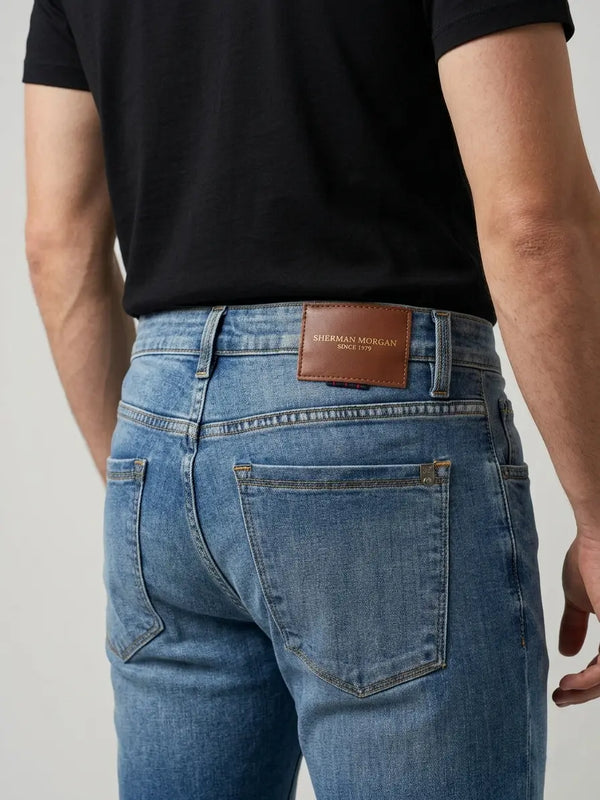 Vista posterior de jeans para hombre con etiqueta de marca en vinipiel café, placa metálica distintiva y bolsillos lisos.