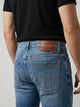 Vista posterior de jeans para hombre con etiqueta de marca en vinipiel café, placa metálica distintiva y bolsillos lisos.