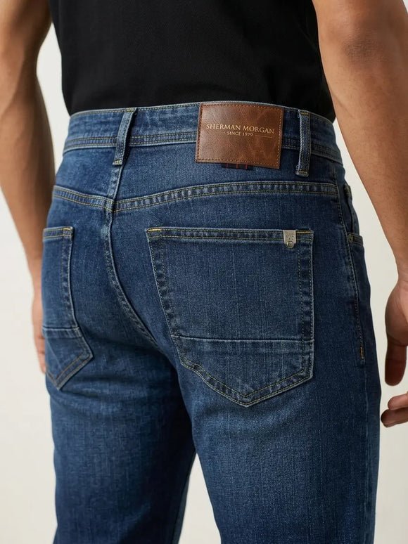 Vista posterior de jeans para hombre con etiqueta de marca en vinipiel café, placa metálica distintiva y bolsillos con diseño de franja horizontal.