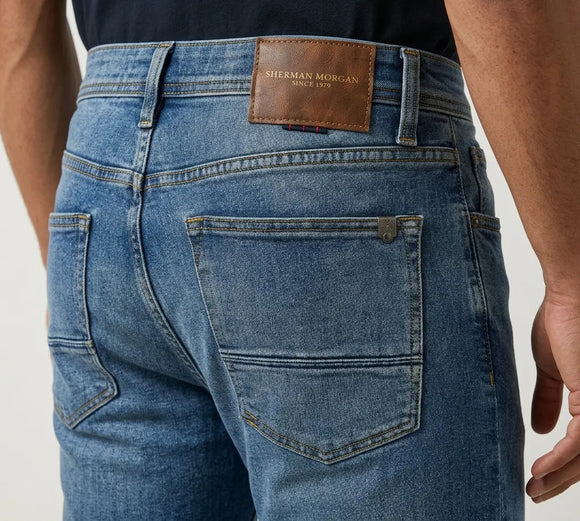 Vista posterior de jeans para hombre con etiqueta de marca en vinipiel café, placa metálica distintiva y bolsillos con diseño de dos franjas horizontales.