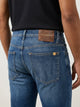 Vista posterior de jeans para hombre con etiqueta de marca en vinipiel café, placa metálica distintiva y bolsillos lisos.
