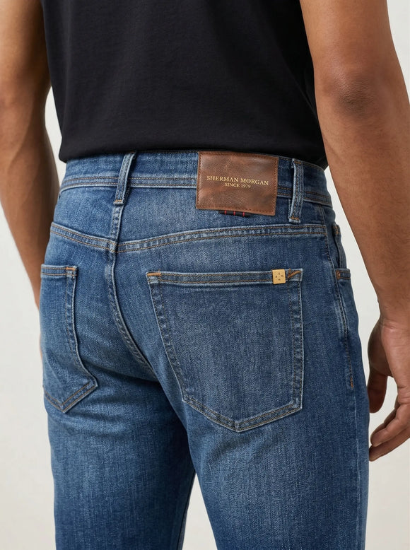 Vista posterior de jeans para hombre con etiqueta de marca en vinipiel café, placa metálica distintiva y bolsillos lisos.