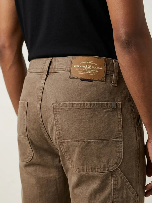 Vista posterior de jeans para hombre con etiqueta de marca en vinipiel café, placa metálica distintiva y bolsillos con diseño de franja horizontal.