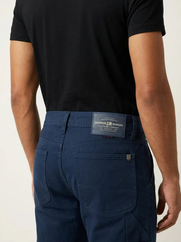Vista posterior de jeans corte carpintero para hombre con etiqueta de marca en vinipiel, placa metálica distintiva y bolsillos lisos.