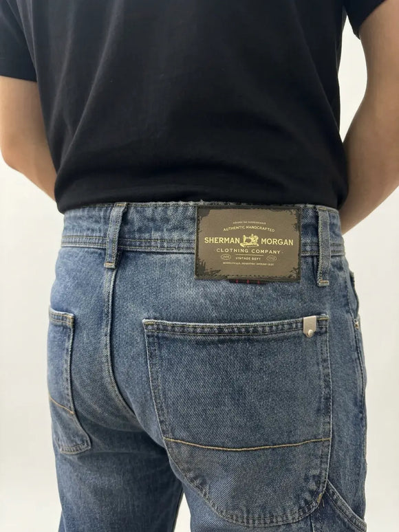 Vista posterior de jeans para hombre con etiqueta de marca en vinipiel, placa metálica distintiva y bolsillos con diseño de franja horizontal.